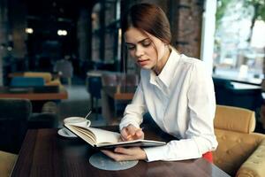 a woman in a shirt with a notebook and a pen in her hand sits at a table in a restaurant on an upholstered chair photo