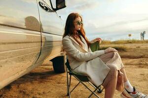 beautiful woman sitting on a chair in a field wearing dark glasses near the car endless field photo