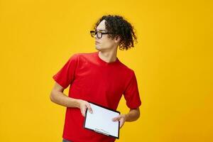 Confused myopic young student man in red t-shirt funny eyewear holds tablet folder with study notes looks aside posing isolated on over yellow background. Free place for ad. Education College concept photo