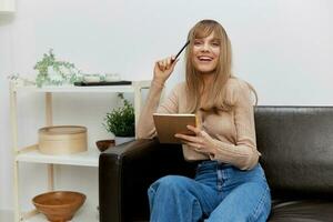 Enjoyed blonde student lady with copybook smiles at camera sitting in sofa couch at modern living room interior. Female client have shrink appointment in a psychological help office. Copy space photo