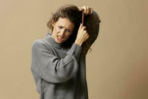 Problematic unruly damaged hair concept. Tormented curly beautiful woman in gray casual sweater with hairbrush pulls tangled hair looks down posing isolated on over beige pastel background. Copy space photo