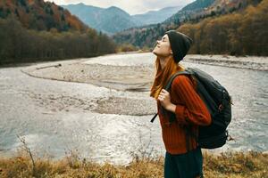 mujer en un rojo suéter y gorra con un mochila en su espalda en el montañas cerca el río en naturaleza foto