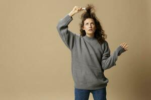 Funny thoughtful curly beautiful female in gray casual sweater with hairbrush comb pulls curls up posing isolated on over beige pastel background. Problematic unruly damaged hair concept. Copy spaceFunny thoughtful curly beautiful female in gray casual sw photo