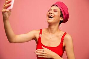 un joven mujer blogger con de colores rosado pelo y un corto Corte de pelo toma un imagen de sí misma en el teléfono y retransmisiones un sonrisa en elegante ropa y un sombrero en un rosado antecedentes monocromo estilo foto