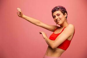 Moda retrato de un mujer con un corto Corte de pelo de púrpura color y un sonrisa con dientes en un rojo parte superior en un rosado antecedentes felicidad foto