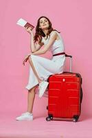 pretty woman in white dress wearing hat red suitcase passports plane tickets pink background photo