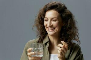 Laughing cute curly beautiful woman in casual khaki shirt hold glass of water smiles looks aside posing isolated on over gray blue background. Healthy lifestyle. Water is Life concept. Copy space photo