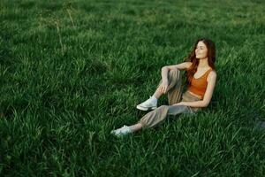 The redheaded woman sits in the park on the green grass wearing an orange top, green pants, and sneakers and looks out at the setting summer sun photo