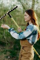 mujer hermosamente sonriente granjero en trabajo ropa y delantal trabajando al aire libre en naturaleza y participación un rastrillo a reunir césped foto