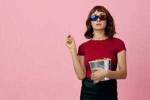 Confused concentrated cute redhead lady in red t-shirt 3D glasses eat popcorn watch movie posing isolated on over pink studio background. Copy space Banner. Fashion Cinema concept. Entertainment offer photo