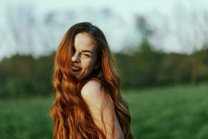 un joven mujer sonrisas y mira dentro el cámara con su largo, rojo, ondulado, brillante pelo en un parque con verde césped en el verano puesta de sol. el concepto de sano viviendo, belleza y pelo cuidado. foto