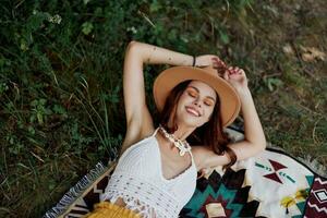 un mujer en Respetuoso del medio ambiente ropa en un hippie Mira es acostado en un de colores tartán sonriente y mirando a un otoño puesta de sol en naturaleza en el parque. estilo de vida en un viaje de armonía con el mundo foto