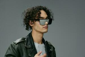 Stylish tanned curly man leather jacket mirror trendy eyewear looks aside posing isolated on gray studio background. Cool fashion offer. Huge Seasonal Sale New Collection concept. Copy space for ad photo
