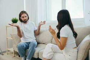 A man and a woman sit on the couch at home in white T-shirts and chatting scandalously do not understand each other. A quarrel in the family of two spouses and aggression photo