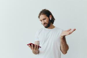 A man with a beard blogger in a white T-shirt with a phone and wireless headphones looks into the phone and spreads his hands to the side on a white background isolated copy space photo