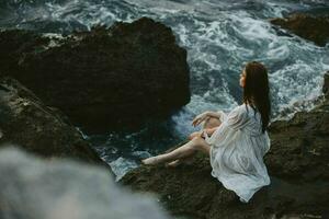 a woman with wet hair in a white dress sits on a cliff barefoot unaltered photo