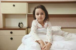 retrato de un caucásico hermosa niño niña en pijama, sentado en el cama a su acogedor dormitorio foto