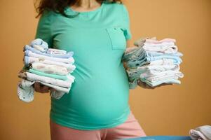 Selective focus on big belly of a pregnant woman, holding folded ironed clean baby clothes over yellow orange background photo