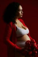 Beautiful multi ethnic pregnant woman holding a bunch of red tulips, posing bare belly over dark red background photo