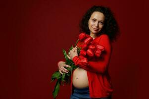 multi étnico hermosa embarazada mujer con un linda ramo de flores de tulipanes, posando desnudo barriga en aislado rojo color antecedentes. foto