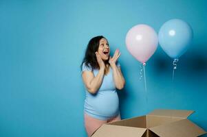 Pregnant woman expressing happy emotions at gender reveal party, blue pink balloons flying out from box - awaiting twins photo