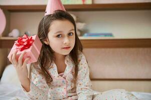 Lovely little child girl shaking pink gift box, trying to guess what is inside it, sitting on the bed in her bedroom photo