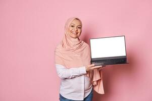 Smiling pregnant Arab Muslim woman in pink hijab, holding laptop with white blank digital screen. Mockup for mobile app photo