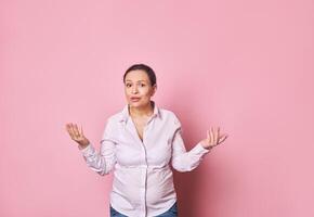 Pregnant woman asking questions, looking perplexed and expressing misunderstanding, posing with hands palms up on pink photo