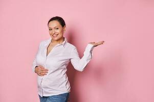 Happy pregnant woman touching gently her belly, smiling at camera, holding imaginary copy ad space on her hand palm up photo
