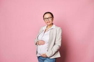 Beautiful middle aged woman touches her tummy, enjoying first baby kicks, expecting the maternity leave, pink background photo