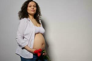 encantador latín americano embarazada mujer en 6 6 mes el embarazo, participación rojo tulipán flor en su desnudo vientre, blanco antecedentes foto