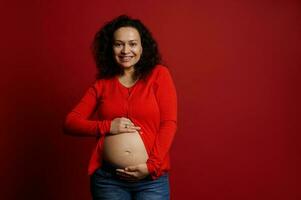 Smiling positive pregnant pretty woman caressing her belly, looking at camera, isolated on red background. Pregnancy photo