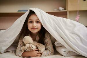 encantador pequeño niño niña en pijama, abrazando su felpa juguete oveja, acostado en su cama debajo cómodo suave blanco foto