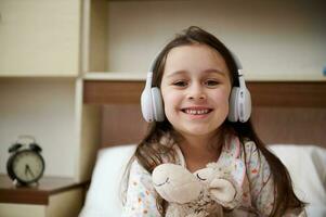 Happy little girl listens to music on headphones, hugs her plush toy and smiles looking at camera, sitting on her bed photo