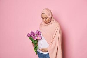 Beautiful Muslim woman with a bouquet of tulips, touching her pregnant belly, isolated on pink background. Copy space photo