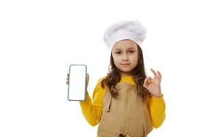 pequeño chica, cocinero confitero, gestos con Okay firmar y muestra a cámara un teléfono inteligente con blanco blanco digital pantalla foto