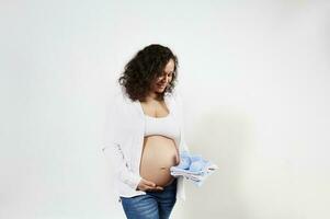 Smiling young charming pregnant woman admiring clothes for newborn baby, gently stroking her naked tummy, white backdrop photo