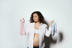 Perplexed pregnant woman, expecting baby, holding blue and pink newborn clothes, guessing the gender of her future child photo