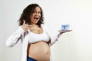 Excited pregnant woman smiles broadly looking at camera, points at blue baby booties in her hand, on white background photo