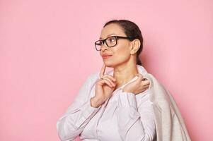 Multi-ethnic pretty woman in trendy glasses, putting her finger on her lips, thoughtfully looking aside on pink backdrop photo