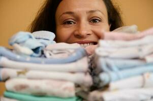 Close-up happy woman smiling looking at camera through a stack of ironed laundered clean newborn clothes photo