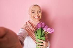 sonriente musulmán mujer en rosado hiyab, hace un autofoto, posando con un ramo de flores de púrpura tulipanes en aislado rosado antecedentes foto