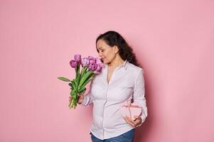 Beautiful pregnant woman sniffing bouquet of tulips, holding a heart shaped gift box with happy present for Mother's Day photo