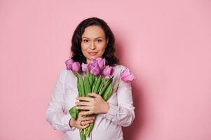 Confident beautiful woman with a bouquet of purple tulips, smiling looking at camera over isolated pink background photo