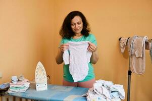 Beautiful pregnant woman, expectant mom holding a laundered baby bodysuit, irons crumpled newborn clothes at home. photo