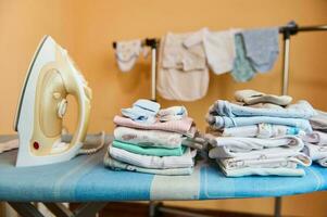 Clean laundered baby clothes ironed and folded in stack near steam iron on an ironing board in dressing room or laundry photo