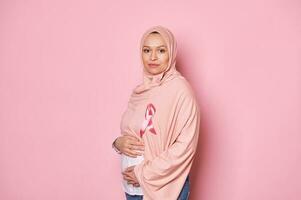 Confident young Middle-Eastern Muslim pregnant woman in pink hijab, wearing a satin awareness ribbon, isolated on pink photo