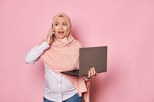 trabajando embarazada árabe musulmán mujer en hiyab, hablando en célula teléfono, participación computadora portátil, teletrabajo el embarazo y carrera foto