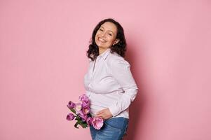 Happy pregnant woman smiling with beautiful toothy smile, posing with a bouquet of tulips, isolated on pink background photo