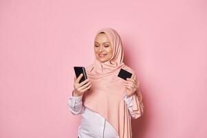 encantador musulmán mujer en rosado hiyab, participación banco crédito tarjeta y teléfono inteligente, hace pagos vía Internet móvil bancario foto